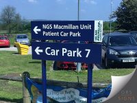 A dual-mounted directional sign, fixed on blue posts in an external hospital car park. The upper sign features a white left-pointing arrow and the text “NGS Macmillan Unit Patient Car Park” in white lettering. A vertical green panel on the right reads “WE ARE MACMILLAN CANCER SUPPORT.” The lower sign displays a white left-pointing arrow and the text “Car Park 7.” Parked vehicles, grass, and a distant rural landscape are visible in the background. The signs provide clear wayfinding information for visitors travelling to the Macmillan Unit and car park facilities.