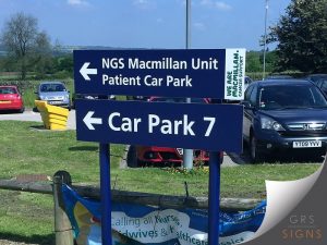 A dual-mounted directional sign fixed on blue posts in an external hospital car park for the NGS Macmillan Unit.