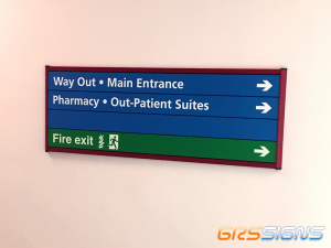 A wayfinding slatz directory wall-mounted sign, showing directions for the way out, the main entrance, the pharmacy, out-patient suites and the fire exit. Majority of the interchangeable slatz are in the NHS' blue, with the fire exit slat being in safety green.