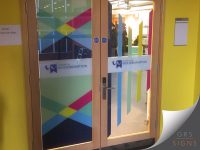 Glass entrance doors featuring vibrant University of Wolverhampton branded window graphics. The design incorporates colourful intersecting geometric shapes in shades of blue, green, pink, and yellow, alongside the University of Wolverhampton logo and text applied across a frosted manifestation band. Additional vertical coloured graphic elements are visible on the glass panels, creating a bright and contemporary entrance feature. The decorative vinyl graphics enhance privacy while reinforcing the university's visual identity and adding colour to the interior space.