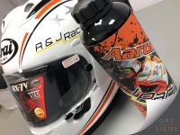 Customised motorsport merchandise featuring a white Arai motorcycle helmet and matching branded drinks bottle. The helmet features a clean white base with black and orange racing stripes, silver star graphics, and personalised A & J Racing branding in black and yellow. The black drinks bottle is wrapped with a vibrant orange, black, and white design incorporating rider imagery, splatter-effect graphics, and prominent Arai branding.