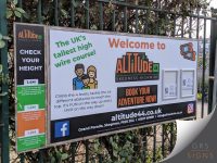 Large roadside welcome and information sign, attached to fence railings for Altitude 44 Skegness Highwire, mounted to perimeter fencing. The design features a predominantly white background with bold orange, green, and black branding, including the Altitude 44 logo and the message "Welcome to Altitude 44". A colourful height-check panel on the left helps visitors determine which high-wire courses they can access, while the main sign includes illustrated graphics, booking information, social media icons, and contact details.