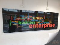 Large acrylic wall panel featuring a colourful business-themed word cloud, mounted to an interior wall using metal stand-off fixings. The design is printed onto a glossy black background and displays key business-related terms in a variety of bright colours, including "enterprise", "business", "corporate", "office", "professional", "strategy", "finance", and "development". The largest words are highlighted in bold red, green, and orange, creating strong visual impact against the dark background.