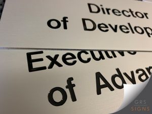 Close-up of a paint-filled engraved brushed metal sign featuring black lettering recessed into a satin silver metallic surface. The sign displays job title information, with the engraved text filled in black to create a sharp contrast against the brushed metal finish. The contemporary design is ideal for office door signs, directories, and corporate wayfinding applications.