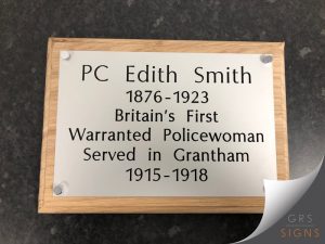 Commemorative plaque honouring PC Edith Smith (1876-1923), Britain's first warranted policewoman, who served in Grantham from 1915 to 1918. The plaque features black engraved lettering on a white panel mounted to a handcrafted oak-effect wooden backing board. Clear acrylic stand-off fixings at each corner give the plaque a contemporary floating appearance, while the natural wood frame provides a warm contrast to the clean white surface and crisp black text.