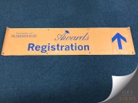Orange University of Huddersfield Awards Registration banner featuring the university logo, elegant script-style "Awards" lettering, and large blue text reading "Registration". A bold blue directional arrow points upwards on the right-hand side, guiding visitors towards the registration area. The banner is finished with reinforced eyelets for hanging and uses a simple, high-contrast design to provide clear event wayfinding.