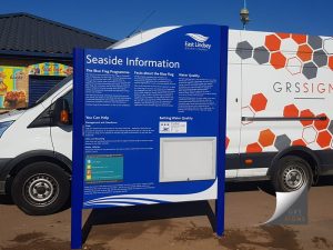 Dual-post, freestanding seaside information sign installed outdoors, featuring a blue and white design with information about the Blue Flag Programme, water quality, beach management, and bathing water quality. The sign displays East Lindsey District Council branding at the top and contains multiple sections of explanatory text, icons, and notices. A lockable display case is incorporated into the lower section of the sign. The sign is supported by two blue posts and is positioned beside a white commercial van displaying GRS Signs branding and an orange and grey hexagonal graphic pattern.