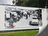 Large-scale external wall graphic at Cadwell Park, featuring a historic black-and-white photograph mounted to the exterior of a building. The image shows a busy race paddock scene with classic motorcycles, cars, support vehicles, and spectators gathered around the circuit during an earlier era of motorsport. The Cadwell Park logo appears in the lower corner, with its distinctive red emblem adding a splash of colour to the otherwise monochrome design. Installed as a prominent feature on the building façade, the graphic celebrates the circuit's heritage and creates a striking visual focal point for visitors.
