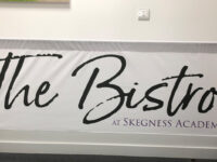 Freestanding café barrier banner for The Bistro at Skegness Academy, mounted between black barrier posts. The banner features a clean white background with "The Bistro" displayed prominently in large black handwritten-style script, creating an elegant and contemporary appearance. Beneath, "At Skegness Academy" is shown in smaller purple lettering, providing a subtle contrast to the monochrome design.