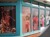 Series of motivational window graphics applied to a school or sports facility, featuring full-height photographs of well-known athletes and inspirational quotes printed across the glazing. The colourful display includes sporting imagery from basketball, athletics, football, golf, and other disciplines, with each panel showcasing a different quote focused on teamwork, preparation, perseverance, and achievement.
