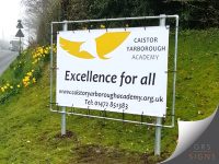 Large outdoor banner for Caistor Yarborough Academy, mounted within a metal frame beside a grassed area and secured via reinforced eyelets and bungee cords. The banner features the academy's yellow and white bird logo alongside the school name in black lettering. Prominently displayed across the centre is the slogan "Excellence for all", with the academy website and telephone number shown below. The clean white background and bold typography create a clear and welcoming sign that promotes the school's identity and values.