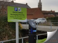 Freestanding electric vehicle charging point sign mounted on a grey post, featuring a bright green background with black text and a blue parking symbol. The sign reads "Electric Vehicle Recharging Point Bay" and "Parking only permitted whilst actively charging", accompanied by an icon showing a car connected to a charging cable. Curved white graphic accents along the bottom add visual interest, while the high-contrast green, black, and white colour scheme ensures the sign is clear, modern, and easily visible within the car park environment.