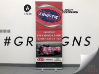 Freestanding pull-up banner advertising Team Christie Racing - featuring a bold red and silver graphic background with a large blue oval logo at the top. Prominent white lettering reads "World Championship Sidecar Team", positioned above an action photograph of a racing sidecar motorcycle carrying the number 34 on a racetrack. Sponsor logos are displayed along the lower section, while GRS Signs branding appears at the base of the banner.