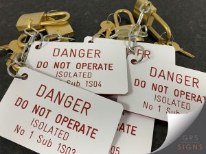 Selection of lockout/tagout safety tags attached to brass padlocks and safety chains, displayed on a dark work surface. The engraved white tags feature high-contrast red lettering reading "DANGER - DO NOT OPERATE", followed by isolation point information and identification numbers. The tags are manufactured from durable acrylic with red engraved text and edges, while the brass padlocks and chains demonstrate their use within a lock-off safety system.