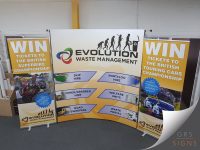 Portable exhibition display stand for Evolution Waste Management, consisting of a curved pop-up backdrop flanked by two roll-up banner stands. The central display features the company logo, the text "Evolution Waste Management", and graphics illustrating services including Skip Hire, Tipper/Grabber Hire, Road Sweepers, Portaloo Hire, Welfare Units, and Waste Portal. The two yellow banners on either side promote competitions to win tickets to the British Superbike Championship and win tickets to the British Touring Car Championship, accompanied by motorsport photography.