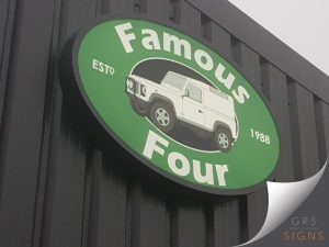 Large oval projecting shop sign for Famous Four, mounted to a dark-clad exterior wall. The sign features a vibrant green background with bold white lettering and a central illustration of a classic white Land Rover set within a black oval. Smaller text reading "ESTD" and "1988" appears on either side of the vehicle graphic, highlighting the company's heritage. The simple colour palette, strong contrast, and vehicle-focused design create an eye-catching sign that clearly reflects the business's connection to Land Rover vehicles and specialist automotive services.