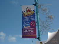 Vertical lamp post banner promoting Fantasy Island, featuring a vibrant purple background with the attraction's colourful logo at the top. The banner highlights attractions including rides for all ages, family fun, live entertainment, and a pay-as-you-ride option. Additional promotional text describes Fantasy Island as the home of Europe's largest seven-day market, with references to fashion, household goods, beauty products, food, drink, and hobbies. Mounted to a turquoise lamp post against a blue sky, the banner uses bold colours and eye-catching graphics to advertise the leisure destination.