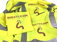 Collection of high-visibility yellow workwear garments branded for Burley & Elsom Electrical. The image shows folded and stacked hi-vis jackets featuring reflective silver safety strips and the company's logo, which incorporates a stylised electrical cable and plug graphic. The branding is printed in brown and grey on the front and back of the garments, demonstrating customised embroidered or printed workwear designed to promote a professional company image while maintaining workplace visibility and safety.