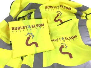 Collection of high-visibility yellow workwear garments branded for Burley & Elsom Electrical. The image shows folded and stacked hi-vis jackets featuring reflective silver safety strips and the company's logo, which incorporates a stylised electrical cable and plug graphic. The branding is printed in brown and grey on the front and back of the garments, demonstrating customised embroidered or printed workwear designed to promote a professional company image while maintaining workplace visibility and safety.