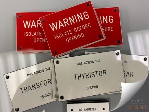 Selection of engraved industrial identification and warning signs manufactured from acrylic. The signs feature a high-contrast colour scheme, including red panels with white engraved text reading "WARNING - ISOLATE BEFORE OPENING" and white panels with black engraved text identifying equipment sections such as "THYRISTOR SECTION" and "TRANSFORMER SECTION". Each sign is pre-drilled in the corners for secure fixing to machinery, control panels, and electrical equipment. The durable engraved construction and clear typography ensure long-lasting identification and safety communication within industrial and engineering environments.