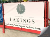 Large PVC banner for Lakings, mounted within a red metal frame. The banner features a clean white background with the Lakings logo and branding in muted green-grey tones, alongside the text "Est. 1908" and "Butchers, Bakers & Delicatessen". A promotional message across the bottom reads "Fresh Meat, Pies & Pastries Available Instore". Secured with eyelets and bungee cords, the banner uses elegant typography and a minimalist design to create a professional and welcoming retail display.