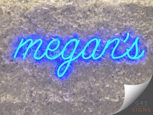 Blue neon-style lettering in flowing handwritten script, reading "megan's". The illuminated sign is mounted against a textured floral backdrop made up of densely arranged white artificial petals, creating a striking contrast between the vibrant blue glow and the soft decorative background. Serving as both a feature wall and an eye-catching branding display, the installation creates a stylish focal point within the interior space.