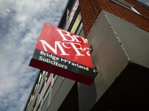 An external modular sign for Bridge McFarland Solicitors. It is a mounted hanging sign featuring the company's colours (deep red, black, and white text)