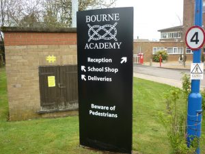Modular sign, black with white text, for Bourne Academy. The sign features directional signage pointing to reception, the school shop, and area for deliveries. It also states to beware of pedestrians.