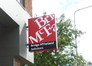An external modular sign for Bridge McFarland Solicitors. It is a mounted hanging sign featuring the company's colours (deep red, black, and white text)