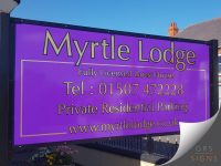 Large freestanding entrance sign for Myrtle Lodge, mounted between two black posts. The sign features a vibrant purple background with elegant gold-effect lettering and border detailing. The business name is displayed prominently across the top, followed by "Fully Licensed Guest House", contact details, and "Private Residential Parking" information. The rich purple and gold colour combination creates a premium, traditional appearance, while the simple layout and oversized typography ensure excellent visibility from the roadside. The sign's black frame and dual-post mounting provide a sturdy and professional finish.