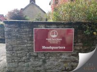 Wall-mounted wayfinding sign for the North York Moors National Park Authority Headquarters, fixed to a traditional stone wall. The sign features a rich burgundy background with white lettering and the National Park Authority logo positioned above the word "Headquarters". A white border frames the sign, creating a strong contrast against the natural stone backdrop.