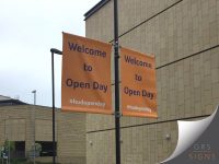 Pair of orange lamp post, double-sided banners, welcoming visitors to a University of Huddersfield Open Day. Mounted back-to-back on a black streetlight pole, the banners feature bold blue text reading "Welcome to Open Day" with the hashtag #hudopenday displayed beneath. Set against the backdrop of a modern university building, the banners provide clear event branding and wayfinding for prospective students and visitors attending the open day.