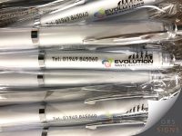 Close-up of a collection of branded promotional pens for Evolution Waste Management, displayed in clear packaging. The white pens feature the company logo, contact telephone number, and an evolution-themed graphic showing silhouettes progressing towards a figure disposing of waste. Chrome trim and clear pen clips add a professional finish, creating a practical and eye-catching promotional giveaway item.