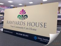 Large entrance sign panel for Rawyards House, a community care home operated by Abbey Healthcare. The sign features a warm cream background with elegant dark blue lettering displaying "RAWYARDS HOUSE" and the tagline "Your Community Care Home" beneath. Above the text, a decorative thistle-inspired logo combines shades of purple, green, and dark blue, adding colour and visual interest. A dark blue footer panel runs along the bottom of the sign, containing the Abbey Healthcare logo, website address, and contact telephone number in white text.