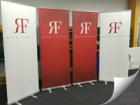Set of freestanding pull-up banner stands branded for Roar Fitness, arranged side by side in an indoor setting. The banners feature a clean, minimalist design, with alternating red and white backgrounds that create a bold yet sophisticated contrast. A large mirrored "RF" monogram logo is positioned centrally above the Roar Fitness name, using elegant typography to reinforce the brand's premium image. The uncluttered layout, limited colour palette, and strong use of negative space give the banners a modern, high-end appearance, making them ideal for exhibitions, fitness events, and promotional displays.
