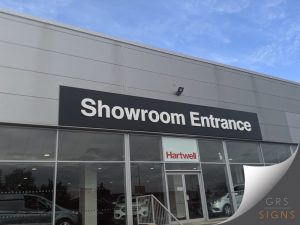 Large showroom entrance sign mounted above the glazed entrance of a Hartwell dealership. The sign features a wide black fascia with bold white three-dimensional lettering reading "Showroom Entrance", creating a strong contrast for excellent visibility. Beneath the sign, the entrance doors and floor-to-ceiling glazing display the Hartwell brand name in red, while the modern grey cladding and clean architectural lines give the building a contemporary, professional appearance. The simple monochrome design ensures the entrance is clearly identifiable and easy for visitors to locate.