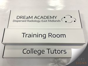Modular slatz sign system for DREaM Academy (Dispersed Radiology East Midlands), featuring a white header panel with the academy logo and branding in black and blue. Beneath, interchangeable aluminium slats display the room names "Training Room" and "College Tutors" in large black lettering. The clean white finish, simple typography, and replaceable slat design create a professional and flexible wayfinding solution, allowing room information to be updated easily within healthcare, education, and office environments.