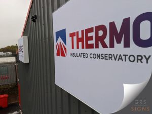 Large wall-mounted sign for Thermotec, fixed to the exterior of a grey corrugated-clad industrial building. The sign features a clean white background with the Thermotec logo in red, blue, and white, alongside bold gradient lettering displaying the company name. Beneath, black text reads "Insulated Conservatory Roof Systems". Mounted on stand-off fixings, the sign projects slightly from the building façade, creating a professional and modern appearance. The simple white background, bold branding, and contrasting colours ensure strong visibility and clear identification of the business premises.