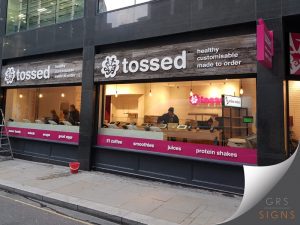 Modern storefront signage for Tossed, featuring a rustic wood-effect fascia with large white 3D/raised lettering and the tagline "healthy, customisable, made to order". A bright pink projecting sign and matching window graphics reinforce the brand's vibrant identity, while a pink band across the shopfront window highlights products including coffee, smoothies, juices, and protein shakes. The design combines natural wood textures with bold white and pink branding, creating a fresh, contemporary appearance that reflects the company's healthy food and drink offering.