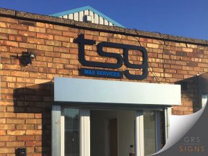 External building sign for TSG M&E Services, featuring bold, 3D black lettering - mounted on a brick façade above a building's entrance. The stylised "tsg" logo is prominently displayed, with "M&E SERVICES" shown below in bright blue capital letters on a black panel.