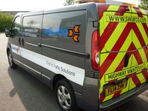 A grey van/safety vehicle, featuring reflective vinyl vehicle livery on the back. Logo is displayed prominently on the side along with relevant company information.