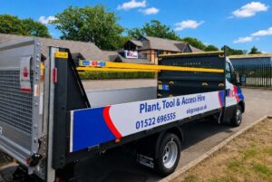 Branded flatbed hire vehicle parked outside commercial premises, featuring side-mounted storage racks carrying long yellow beams. The vehicle displays VIC Group plant, tool and access hire branding, contact details, and website information, with a clear blue sky and industrial buildings in the background.