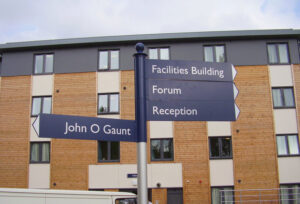 A fingerpost sign aiding with wayfinding, pointing out directions for reception, forum, facilities building and John O Gaunt in navy blue with white text.