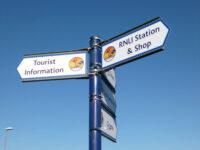 A cantilever fingerpost sign in Mablethorpe, aiding with wayfinding for RNLI's station & shop, tourist information, the cinema, toilets and the local seal sanctuary. Post is navy blue, signs are white with a Mablethorpe mermaid logo and blue outline.